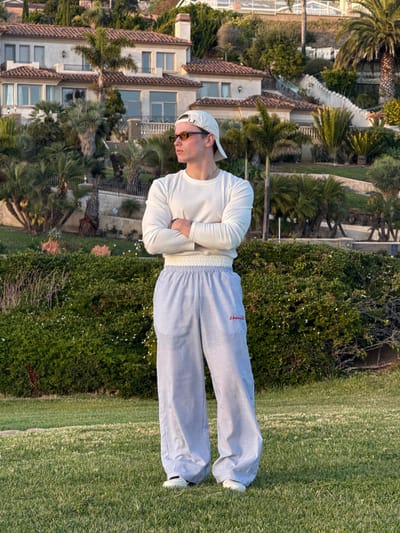 A medium shot, captured from a high-angle perspective, presents a man standing on manicured green grass adjacent to a robust, verdant hedge. The location is identifiable as Rancho Palos Verdes, United States of America, based on the architecture and flora visible in the background.
The man, positioned centrally in the frame, is clad in a white, long-sleeved, striped top and horizontally striped blue and white pants, with his arms folded across his chest. White sneakers are visible on his feet. He is wearing a baseball cap and sunglasses. The surrounding environment includes residential buildings with red tile roofs, indicative of the architectural style prevalent in the region, complemented by palm trees and other vegetation.