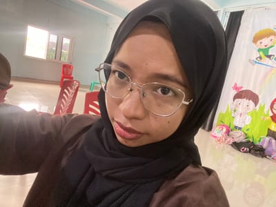The image shows a close-up, selfie-style shot of a young woman wearing a black hijab and glasses. She is wearing a brown shirt. The background appears to be an indoor space, possibly a classroom or a community hall, with a window on the left showing some greenery outside. Red chairs are visible in the middle ground, and a colorful mural or banner with cartoon characters is present on the right. Several backpacks are scattered on the floor near the mural. The lighting suggests it is daytime. The location is specified as Tanjung Gading, Indonesia.