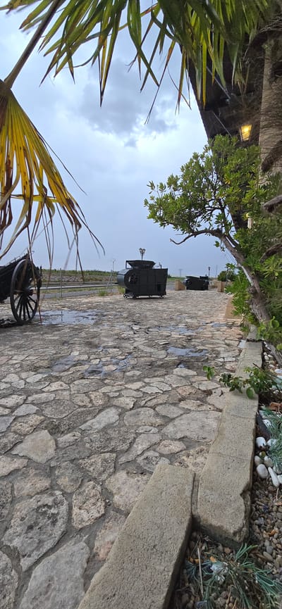 The image depicts an outdoor scene at Venta de la Romera, Spain. The foreground is a stone-paved area with puddles of water, suggesting recent rain. To the left, a portion of an old wagon wheel is visible. In the mid-ground, there are two dark, industrial-looking machines on wheels, resembling old farming equipment or possibly some type of generator or processing unit. Behind these machines, a metal fence runs horizontally, with a hint of greenery and possibly cultivated fields beyond. To the right, a portion of a building with a wooden roof and an outdoor light fixture is visible, along with a lush green tree. The sky is overcast with grey clouds. The overall impression is of an outdoor exhibit or historical site, possibly at an inn or rural establishment.