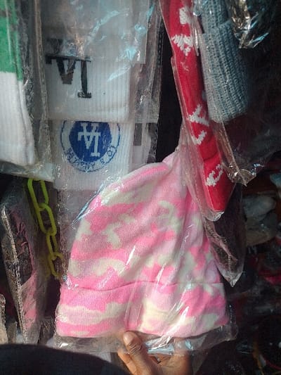 The image presents a close-up view of various packaged clothing accessories, observed within what appears to be a market setting in Ajah, Nigeria. In the lower-center, a dark-skinned hand holds a pink and white camouflage-patterned beanie, which is sealed within a clear plastic bag. To the upper-left, several white items, identifiable as socks or wristbands, are individually packaged in clear plastic. One visible item displays the black letters "LA," and another beneath it features a blue circular emblem with "LA" and additional text. To the upper-right, a red beanie adorned with white patterns and a grey ribbed beanie are also enclosed in clear plastic bags. A bright yellow plastic chain or similar item is partially visible on the far left. The background is dark and indistinct, consistent with a dense retail display or stall.