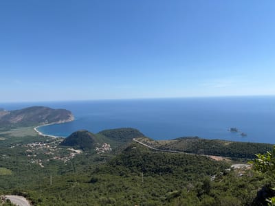 The image shows a panoramic view from a high vantage point overlooking the Adriatic Sea and a coastal landscape. In the distance, a large, verdant mountain juts out into the sea, with a curved coastline and a beach visible. A small town or village with red-tiled roofs is nestled among the green hills in the mid-ground. Roads wind through the hilly terrain, with one road prominently featured leading towards the sea. Several smaller, rocky islands or islets are scattered in the blue water further offshore. The sky is a clear, bright blue with no clouds. The foreground is dominated by dense green foliage and trees, suggesting a natural, mountainous environment. The overall impression is one of a scenic coastal region in clear weather, likely Montenegro given the mention of Novoselje, which is a coastal settlement in Montenegro.