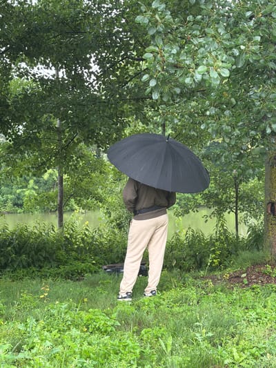 A person stands under a large black umbrella, facing away from the camera. They are wearing a dark green jacket and light beige pants, with black and white sneakers. The person is positioned on a grassy bank, with lush green foliage and trees surrounding them. In the background, a calm body of water, possibly a lake or river, is visible, bordered by more dense greenery. The overall scene suggests an outdoor setting, likely during a light rain, given the umbrella. The location is specified as Nikulskoye, Russia.