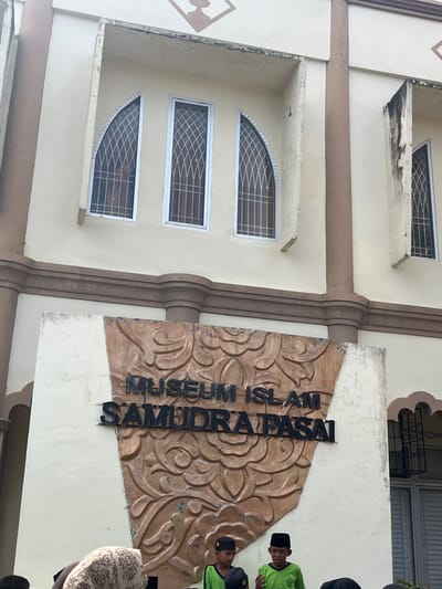 The image shows the facade of the Museum Islam Samudra Pasai, located in Kruengmate, Indonesia. The building is cream-colored with brown trim and features arched windows with metal grilles. A large, decorative sign in the foreground reads "MUSEUM ISLAM SAMUDRA PASAI". Several people, including young boys wearing black peci hats and green shirts, are visible at the bottom of the frame, suggesting a public or educational visit. The overall scene depicts an exterior view of a cultural institution.