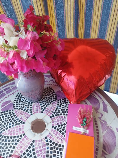 A still life scene is set on a mosaic-tiled table, possibly outdoors given the natural lighting. In the background, a striped curtain with vertical bands of blue, yellow, and pink hangs, suggesting a domestic or decorative setting.

The primary subjects are a vase of vibrant pink and white bougainvillea flowers, a large red heart-shaped balloon, and a small gift card with a decorative dried flower arrangement. The vase is lavender-colored with speckles of red and black, and it holds a generous bunch of bougainvillea. To the right of the vase, the shiny, crinkled surface of the red heart balloon is prominent. In the foreground, a pink card is adorned with a small bundle of dried grass and a few green embellishments. Below this, a small orange box or card is visible. A circular chocolate or candy is placed in the center of a white sunburst design on the mosaic table.

The overall mood is celebratory and thoughtful, perhaps indicating a gift-giving occasion such as Valentine's Day or an anniversary. The bright colors of the flowers and balloon evoke a cheerful atmosphere.

Visible text on the card reads: "Especially For You".

The location context is identified as Al Ahia', Egypt. The time of day appears to be daytime due to the lighting conditions.