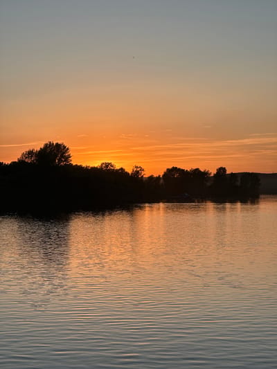 The image depicts a sunset over a body of water, likely a river or lake. The sun is setting on the left side of the frame, casting an orange glow on the sky and reflecting on the water's surface. A line of trees or vegetation is silhouetted against the setting sun, forming a dark barrier between the sky and the water. The water is rippled, and the reflections of the light and trees create a textured pattern. The specific location of Nikolo-Beryozovka, Russia, is not discernible from the visual information alone.