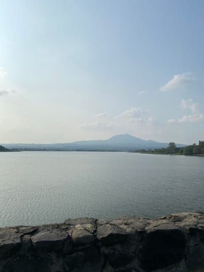 The image depicts a serene natural landscape, likely a body of water such as a lake or reservoir, bordered by a rocky embankment on the right. The water's surface is rippled, suggesting a gentle breeze. In the distance, a chain of mountains fades into a hazy blue silhouette against a pale sky. The sky is mostly clear with a few wispy clouds, indicating fair weather. The light suggests it is either early morning or late afternoon, with the sun casting a soft, diffused glow. The scene is tranquil and peaceful, evoking a sense of calm. The rocky embankment appears to be man-made or enhanced, possibly part of a dam or a constructed viewpoint. The overall impression is one of natural beauty and stillness.