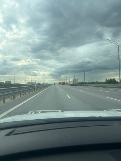 The image captures a view from inside a vehicle driving on a multi-lane highway under a cloudy sky. The highway appears to be an elevated road or a bridge, with metal guardrails lining the right side. Several vehicles, including trucks and cars, are visible on the road ahead and in the distance. On the left side of the highway, there are buildings and trees. The sky is filled with dark, heavy clouds, suggesting impending rain or overcast weather. Streetlights are positioned along the highway. The foreground shows a portion of the vehicle's dashboard and windshield. The scene is likely a highway outside of a city.