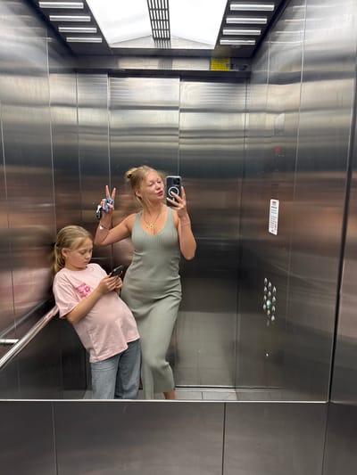 Two females are standing inside a stainless steel elevator cabin. One woman wears a striped olive green dress and the other wears a pink t-shirt and gray jeans. The woman in the dress holds a smartphone and extends her other hand with a peace sign. The other woman is holding a smartphone and looking at it. The location is an elevator, but the city of Ariel in the Palestinian Territories cannot be definitively determined from the image.