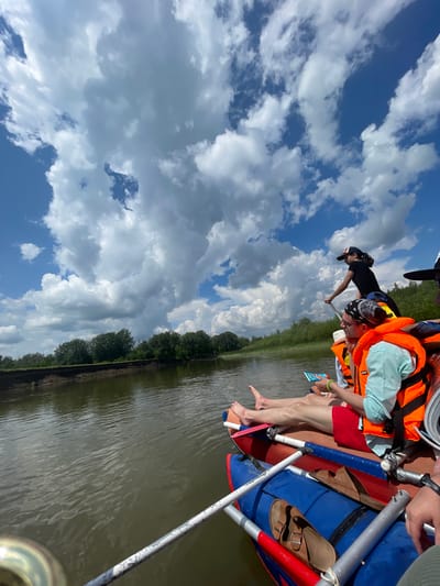 An outdoor, eye-level, medium shot shows a group of people on an inflatable raft on a river under a cloudy sky. The raft, with red, blue, and white accents, carries at least four people. Two individuals in orange life vests are visible in the foreground. One person, wearing a blue shirt and red shorts, has their legs extended, and a book rests on their lap. Another person, also in an orange life vest, has their head turned away from the camera. A person in the background, wearing a black t-shirt and a black baseball cap, holds a paddle. The river, reflecting the sky, occupies the lower left of the frame, with a wooded embankment on the far side. The sky is filled with large, dramatic white and gray clouds. The scene appears to be located in the city of Nezhinka, Russia.