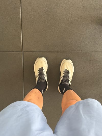 In Montuïri, Spain, a person stands on a dark grey, textured floor composed of interlocking square tiles. They are wearing light-colored athletic shoes with black laces and black socks. The lower half of their legs and thighs are visible, with one leg showing minor abrasions on the skin. They are also wearing light blue shorts. The image is taken from a top-down perspective.