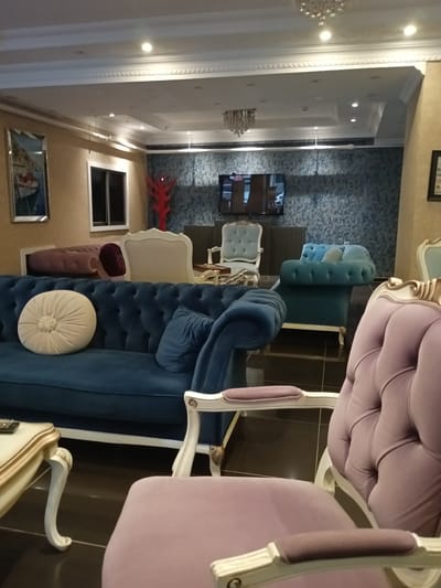 This image shows a well-lit, interior space, likely a hotel lobby or a furniture showroom, in Kırıkkale, Turkey. The room features multiple upholstered seating arrangements in various colors, including a large, dark blue tufted sofa with a round cream cushion and a smaller blue cushion, and a plush purple armchair with a decorative white frame. A light-colored, ornate coffee table with a remote control on its surface is positioned in front of the blue sofa. In the background, there is a light blue tufted sofa, a white ornate chair, and a pinkish tufted sofa. A television is mounted on a wall with patterned wallpaper. The ceiling is adorned with decorative molding and recessed lighting, and a chandelier hangs from the ceiling. On the left wall, there is a framed picture depicting a boat. The flooring appears to be dark tile. There are no people visible in the image.