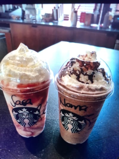 The image depicts two personalized Starbucks Frappuccino beverages positioned on a dark, reflective table surface inside a Starbucks coffee shop located in Soweto, South Africa. The beverage on the left, contained in a clear plastic cup, presents a stratified appearance with a red, possibly strawberry, layer interspersed with white elements, culminating in a substantial topping of white whipped cream beneath a clear plastic dome lid. The name "Sasa" is handwritten in black marker on this cup. Adjacent to it, on the right, is another clear plastic cup holding a brown-toned beverage, indicative of chocolate or coffee, also crowned with white whipped cream and a pattern of dark chocolate drizzle, secured by a clear plastic dome lid. The name "Noma" is handwritten in black marker on this cup. Both cups prominently display the Starbucks siren logo. The background is an out-of-focus view of the coffee shop's service counter, revealing various stainless steel and glass fixtures, bottles, and equipment typical of a commercial beverage preparation area. No individuals are visible in the scene.