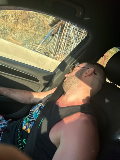 A man wearing a black tank top and glasses is asleep in the passenger seat of a car. He is buckled in. A construction site with scaffolding is visible through the window. The car is located in an urban environment, potentially Agios Pavlos Municipal Unit, Greece.