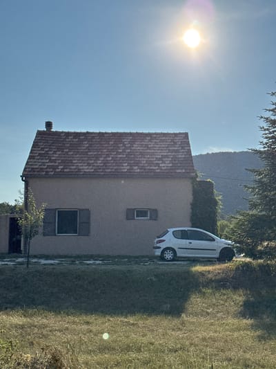 The image depicts a residential scene in Kopito, Montenegro, bathed in bright sunlight. In the foreground, a patch of dry grass and vegetation is visible. Behind this, a low mound of grass leads up to a paved area. A white compact car is parked on this paved surface to the right of a light-colored, two-story house with a red-tiled roof. The house has two windows with brown shutters, one larger and central, the other smaller and to the right. A chimney is visible on the roof. To the left of the house, a thin tree stands next to an open doorway. To the right of the house, a large evergreen tree is present. In the background, a hazy, dark mountain range is visible under a clear blue sky, with the sun prominently positioned in the upper right quadrant, casting a bright glare. The overall impression is of a rural or semi-rural residential property.