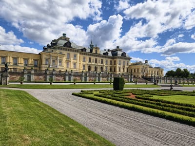 This is an outdoor, eye-level, wide-angle photograph of the exterior of the Drottningholm Palace in Ekerö kommun, Sweden. The palace, painted a pale yellow, is a large, baroque-style building with multiple wings, domes, and classical architectural details. It is set against a bright blue sky filled with large, fluffy white clouds. In the foreground, there is a meticulously maintained garden with manicured green hedges forming geometric patterns, interspersed with patches of reddish-brown soil. A gravel pathway curves through the garden. To the left of the gravel path is a vibrant green lawn. A tall, dark green, rectangular hedge stands prominently in the middle-right of the garden. Behind the garden, a stone wall with decorative balustrades stretches across the width of the palace. Statues and decorative urns are visible along the wall and on the palace's terraced entrance. The overall scene depicts a grand architectural structure surrounded by formal gardens under a sunny, cloudy sky.