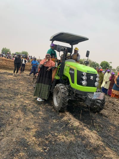 A green WOFAN tractor is parked on a dirt field with black charred grass. A woman wearing a colorful dress and a headscarf is standing next to the tractor, holding a microphone and speaking. Several other people are standing around the tractor, including a woman in a purple headscarf. The tractor is located in a field near a small village with several buildings and trees in the background. The location is a rural area, likely a farm or field near Ringimawa, Nigeria, based on the provided coordinates. The sky is partly cloudy and the sun is shining.