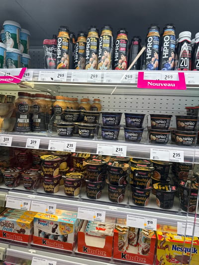The image shows a refrigerated display case in a grocery store, likely a supermarket, in Echternach, Luxembourg. The shelves are filled with various food and drink items, primarily focusing on high-protein products and desserts.

On the top shelf, there are several bottles of "HiPRO" protein drinks in different flavors, including "0% mangue" (0% mango) and "0% d. van biscuit" (0% vanilla biscuit). These bottles are black with prominent "HiPRO" branding and images of men. There are also some "Starbucks Cafe Latte" cups visible on the far left.

The middle shelves are stocked with "HiPRO" yogurt cups, featuring flavors like "Myrtille-Bosses" (Blueberry) and "Beurre de cacahuète-banane" (Peanut butter-banana). Below these, there are bottles of "CAFFE LATTE HIGH PROTEIN COFFEE" and more "HiPRO" protein drinks.

The bottom shelves display a variety of desserts, including "Ehrlmann High Protein Chocolate Topping," "High Protein Vanilla Pudding," and "High Protein Caramel Style Pudding." Further to the right, there are boxes of "Kinder Choco Fresh," "Kinder Pingui," and "Nestle Nesquik Cacao."

Price tags are visible for many of the products, indicating prices in Euros. Some of the labels also show weight and nutritional information. A prominent pink "Nouveau" (New) sign is attached to one of the shelves, highlighting new products. The overall environment is a well-lit, organized retail setting, typical of a supermarket's refrigerated section. No people are visible in the image.