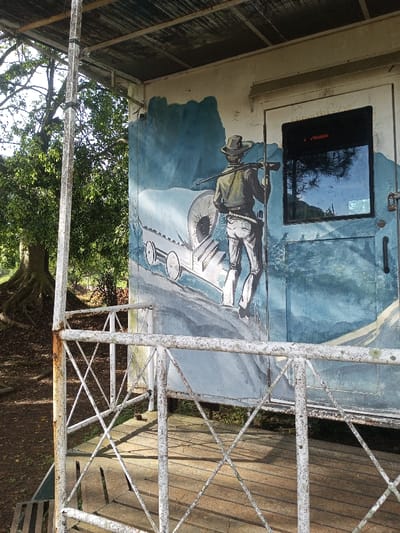 A mural on the side of a building depicts a cowboy leading a covered wagon up a hill. The cowboy wears a hat and carries a rifle over his shoulder. The scene is painted in shades of blue, gray, and white. A window is set into the side of the building next to the mural, reflecting the sky and trees. A weathered white metal railing in the foreground partially obscures the view. A tree is to the left of the structure.
