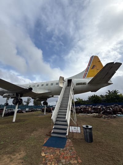 An airplane with stairs leading to the entrance is parked on a grassy field in Kraaifontein, South Africa.  The plane has yellow tail markings with red, white and blue stripes. An "Enter at Own Risk" sign stands near the stairs and a metal trash can is nearby. Several vehicles are parked in the background.  The sky is cloudy.
