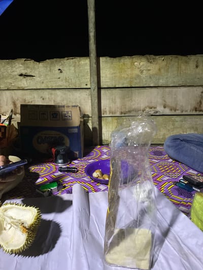The image depicts an outdoor scene at night, possibly in Lamtui, Indonesia, with a wooden plank wall in the background. In the foreground, a durian fruit is partially opened on a white surface. A purple woven mat with a geometric pattern lies on the ground, holding various items. These include a purple bowl with several small, pale yellow objects, a transparent plastic bag containing a pale yellow substance, a black flashlight, a green lighter, and a smartphone. To the right, a person's legs, clad in blue jeans, are visible, suggesting their presence at the scene. A cardboard box labeled "Qatar" is also present. The overall lighting is dim, with shadows cast by the objects.