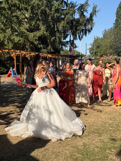 The image depicts an outdoor wedding scene, possibly in a garden or park setting, with a bride in a white gown at the forefront, holding a bouquet. She is facing towards the viewer and appears to be smiling. Behind her, a group of women, presumably bridesmaids or guests, are gathered, some in colorful dresses. The environment is sunny with visible trees and greenery. There is a wooden structure with lights in the background, and a children's slide is visible to the left. The ground is covered with dry grass. The image does not contain any information about the city of Hem, France.