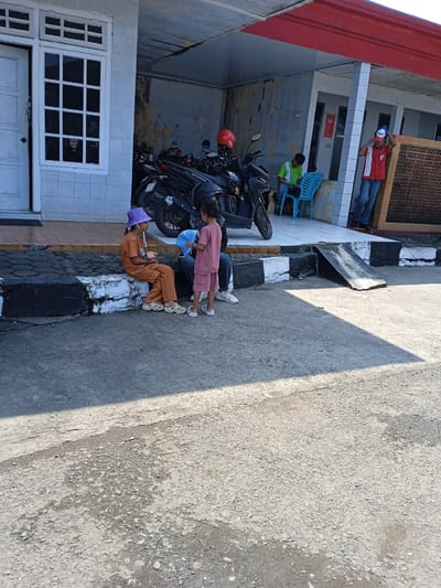 Two young girls sit and stand on a black and white curb near a building in Brangsong, Indonesia. The girl on the left is wearing brown clothing and a purple hat, while the girl on the right is wearing a pink outfit. An adult sits between them. Behind them are parked motorcycles under the building's shelter. Further back, two men are visible near the building's entrance. The area is paved, showing signs of wear and tear. The building has a white door and window to the left and a red roof overhangs the scene.