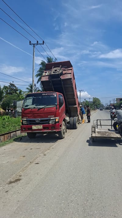 This is a photograph taken outdoors in Beurandang, Indonesia on a bright, sunny day with a partly cloudy sky. The main subject is a red dump truck with its bed tilted upwards, positioned on a paved road. Two men are visible near the rear of the truck. One man, wearing a blue shirt and orange boots, appears to be interacting with the truck's mechanism or the ground beneath it. Another man, in a brown shirt, stands nearby, observing. To the right of the dump truck, a motorcycle with a trailer is parked, and a person is seated on the motorcycle. Further down the road, other vehicles, including cars and motorcycles, are visible in the distance, suggesting ongoing traffic. The environment includes a grassy embankment to the left of the road, lined with vegetation and palm trees. Electrical poles and wires are strung across the scene, typical of urban or semi-urban infrastructure. The road surface is asphalt with some visible patches and minor debris. The overall impression is a street scene with a construction or maintenance activity involving the dump truck.