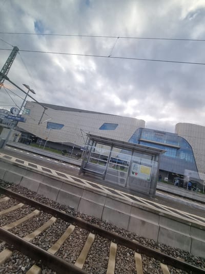 The image depicts a view from a train approaching or departing from Weil am Rhein, Germany. The scene is dominated by the Weil am Rhein railway station, recognizable by its modern architectural design and large glass windows. A bus shelter is visible on the platform. Train tracks run along the right side of the image, partially obscured by ballast. Overhead power lines and their support structures are visible against a cloudy sky. The station building has the name "Weil am Rhein" visible on the facade.