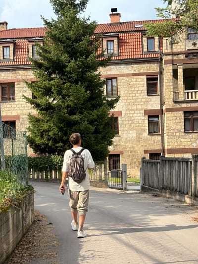 A person stands on a paved path next to a building. The building is made of light-colored stone and has red-tiled roofs. The person is facing away from the camera, wearing a white t-shirt, tan shorts, and white sneakers. They have a dark backpack on their shoulders and are holding a black smartphone in their right hand. A large evergreen tree is positioned in front of the building. The scene appears to be outdoors during the day, with shadows cast on the path, suggesting sunlight. The location context is Jajce, Bosnia and Herzegovina. No visible text is present in the image.