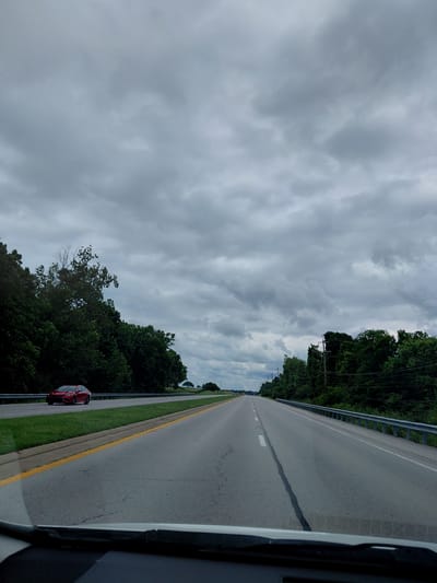 The image depicts a roadside scene under a cloudy sky. The perspective is from inside a vehicle, looking out towards a highway. The highway stretches into the distance, with a central lane marking and white dashes dividing the lanes. A silver guardrail runs along the left side of the road, separating it from a dense line of dark green trees that create a natural barrier. A bright red car is parked on the shoulder of the road, near the guardrail. The sky is filled with a thick layer of grey clouds, suggesting overcast weather, possibly with a chance of rain. The overall scene is serene, with the natural landscape of trees contrasting with the man-made road. There is no visible text in the image. The time of day appears to be daytime, but the overcast conditions limit direct sunlight. The location is identified as Versailles, United States of America.