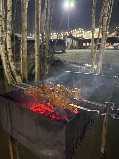 The image displays an outdoor night scene in Vasil'evo, Russia, featuring a metal brazier (mangala) actively cooking shashlyk. Three metal skewers, laden with marinated meat pieces, are positioned over glowing red charcoal embers, emitting visible smoke. The brazier stands on wet ground, possibly shallow standing water, indicated by reflections beneath its legs. In the midground, several slender, light-barked trees, consistent with birches, stand amidst wet, possibly snow-covered ground. Beyond these trees, a paved or dirt area serves as a parking lot, with several dark-colored vehicles parked. Further in the background, illuminated buildings featuring distinct roofs and decorative string lights suggest a recreational facility or restaurant. A brightly lit streetlamp with a greenish light source is visible centrally. In the far background, a sloped terrain is densely dotted with numerous small lights, indicative of a lit ski slope or winter recreation area. No individuals are present in the frame.