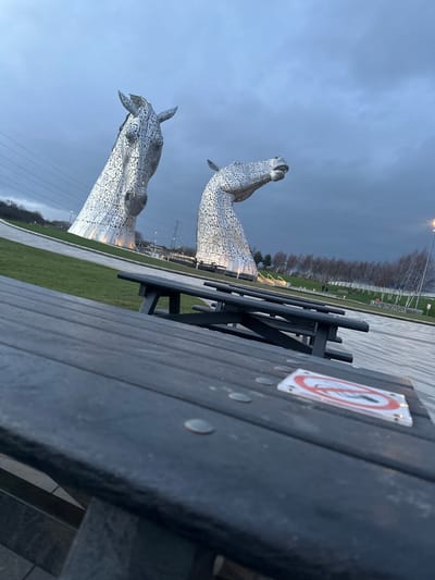 The image displays an outdoor scene featuring The Kelpies, two colossal metallic horse head sculptures, under an overcast, dark grey sky. The location is The Helix Park, Falkirk, Scotland, United Kingdom.
In the immediate foreground, a dark-colored wooden picnic table occupies the lower portion of the frame, angled upwards from left to right. Visible on its slatted surface are several small, round, silver-colored metallic objects, and a white square sticker with a red circular border enclosing a diagonal line through a lit cigarette, indicating a "No Smoking" prohibition.
Behind the picnic table, a paved walkway leads towards the bases of the two horse sculptures. The sculpture on the left, depicted with its head slightly raised and facing left, is partially obscured by the table's perspective. The sculpture on the right presents a horse head looking straight ahead with its mouth slightly open. Both sculptures are composed of numerous polished metal plates and appear illuminated from below.
The midground features green grassy areas and paved pathways surrounding the sculptures. In the background to the left, a grassy slope rises, with power lines and a large metal pylon visible against distant trees. To the right, a row of slender, light-barked trees extends horizontally, bordering what appears to be a canal or water feature, with additional distant structures and a street light also visible. No people are present or identifiable in the scene. There are no explicit actions or interactions depicted.