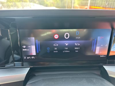 The image shows a close-up view of a car's digital dashboard display. The dashboard is wide and modern, with a dark screen displaying various vehicle information. In the center of the display, the drive mode is indicated as "D" for Drive, with the current speed shown as "0 km/h". A red circular sign with "10" inside suggests a speed limit of 10 km/h.
To the left of the center, a fuel gauge shows the fuel level at "1/2" and the remaining range as "306 km". Below this, the outside temperature is displayed as "47°C" along with a warning triangle.
To the right of the center, there are icons representing different vehicle functions. One icon with a car and a battery symbol indicates a range of "552.1 km", and another shows "549 km" as the total distance. A fuel consumption reading of "7.8 L/100km" is also visible. On the far right, a temperature gauge is displayed with readings at "90" and "50", and a thermometer icon indicates the outside temperature.
The overall setting appears to be inside a vehicle, likely during the day given the bright light filtering from the top of the image, which suggests an outdoor environment. The dashboard's design implies a modern vehicle. There are no people visible in the image. The location context provided is Castiadas, Italy, though no specific visual cues within the dashboard itself confirm this.