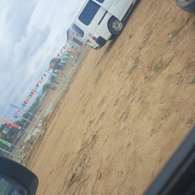 The image, taken in Wuro Ngangandi, Nigeria, depicts an outdoor scene with a white van parked on a dirt surface. A fence with colorful flags and banners strung along it is visible in the background. The flags appear to be triangular and are red, orange, blue, green, and white. Some of the flags have black symbols on them. Beyond the fence, trees and a cloudy sky are visible. The dirt ground is uneven and has scattered dry vegetation and some debris. The van is white and appears to be a panel van with tinted windows. The image is taken from a low angle, possibly from inside another vehicle, as a portion of a dark car interior is visible in the lower left corner of the frame. The overall impression is of an open area, possibly a community space or event venue, in Nigeria.