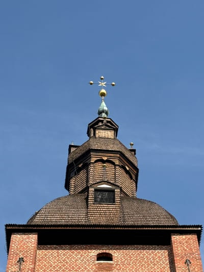 The image shows the upper part of a brick building, likely a church tower, in Askersund, Sweden. The building features a domed roof covered in wooden shingles, transitioning to an octagonal structure also clad in shingles. A smaller, pointed roof section sits atop this, topped with a green spire and a golden weather vane with a cross shape. The sky is clear and blue. The brickwork of the lower visible section of the building is red and laid in a stretcher bond pattern. There are no people or animals visible in the frame.