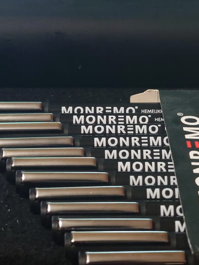 The image shows a close-up, angled view of a set of metal rods, likely tools, arranged in rows. The rods have a metallic sheen and appear to be cylindrical. Each rod is positioned in front of a black surface with white lettering. The visible text on the black surfaces repeats the word "MONREMO" and other partial words or symbols. The arrangement suggests a tool organizer or storage case, with each rod fitting into a designated slot. The overall environment appears to be indoors, with a dark background and diffused lighting, possibly indicating a workshop or storage area. There are no people visible in the image. The image quality is good, allowing for clear observation of the objects and text. The provided location of Sokolohirsk, Ukraine, is noted but not visually discernible from the image itself.