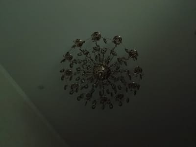 A chandelier hangs from a dark, muted ceiling. The chandelier is intricate, with many crystal-like elements that reflect light. It appears to be made of metal and glass, with a floral or starburst design. The overall lighting is dim, creating a somber atmosphere. The image does not provide any contextual information about the location in Nukus, Uzbekistan.
