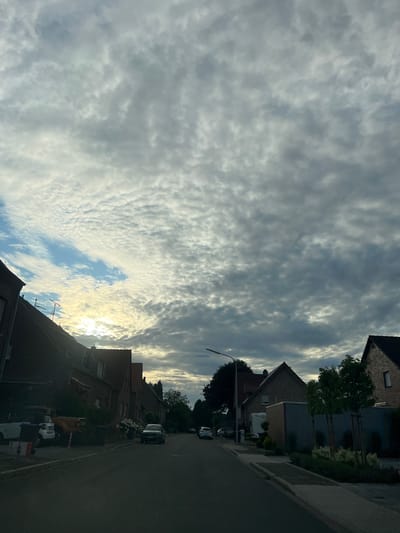 The image depicts a street scene in Heinsberg, Germany, under a cloudy sky. The perspective is from a street, looking down a residential road lined with brick houses. Several cars are parked along the street and on the driveways of the houses. The sky is filled with a textured pattern of gray and white clouds, with patches of blue sky visible and the sun partially obscured by the clouds, casting a soft light. Some houses have antennas on their roofs. Trees and bushes are present in the yards of the houses and along the side of the road. The street itself is paved and appears to be in good condition. The overall atmosphere is calm and residential.