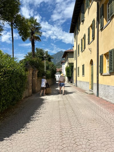 The image captures a sunlit, narrow paved street in Abbadia Lariana, Italy. Two individuals are observed from behind, walking away from the camera. The person on the left wears a white top and patterned skirt or shorts, pulling a large, light-brown wheeled bag or cart. The person on the right wears blue patterned shorts, a light-colored shawl draped over their shoulders, and a light-colored straw hat.
The street surface consists of light-colored pavers arranged in a repeating fan or radial pattern. On the left, a dense green hedge borders a stone wall with an ornate, arched gate. Two tall trees, one distinctly a palm, rise above the hedge line. A streetlamp pole is visible further down the path. On the right, a multi-story building with a light yellow stucco facade presents multiple windows fitted with closed, dark green wooden shutters. A dark green entrance door with the number "42" affixed is visible towards the foreground. The building's base features a grey, textured finish. In the background, distant mountains are visible under a blue sky with scattered white clouds. Strong sunlight casts distinct shadows from the foliage and the pedestrians onto the paved street.