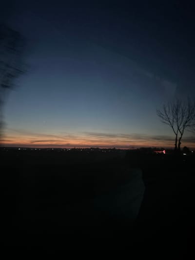 The image depicts an outdoor landscape scene at twilight, observed in or around Senokos, Bulgaria. The sky transitions from a deep indigo at the zenith to shades of blue and then a prominent band of orange and red hues along the horizon, indicative of either dusk or dawn. Faint, elongated cloud formations are visible within the orange-red stratum. A single bright celestial object is present in the upper left quadrant of the sky.
Along the distant horizon, a series of small, bright artificial lights are discernible, signifying a populated area. A distinct, leafless deciduous tree, characterized by its skeletal branches, stands silhouetted against the lighter sky in the right portion of the frame. A blurred, dark, vertical element is partially visible on the far left edge of the image, suggesting nearby vegetation or an obstruction close to the camera. The foreground beneath the horizon lights is obscured in deep shadow, appearing as an indistinct, dark landmass. Translucent reflections are subtly present across the image, particularly as a lighter, rectangular form in the upper right sky and an amorphous shape in the lower-mid foreground, suggesting the photograph was taken through a reflective surface, such as a window. No individuals, direct actions, or interactions are visible within the scene.