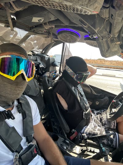 The image displays two individuals inside a utility terrain vehicle (UTV) situated on a paved roadside in Kefar Sava, Israel. The scene, captured from the passenger's perspective as a selfie, features a bright, clear sky overhead and dry vegetation bordering a metal guardrail in the background.

The passenger, on the left, wears a white t-shirt, dark lower garments, a grey balaclava covering the lower face, and large, yellow-framed goggles with a multi-colored reflective lens. They are secured by a black multi-point safety harness.

The driver, on the right, wears a black t-shirt or tank top, patterned shorts, a black head covering, and goggles with "LEATT" visible on the strap. They are also secured by a black multi-point safety harness or vest. The driver's left hand is near the steering wheel.

The UTV's interior is rugged, featuring visible roll cage components, overhead speakers with blue LED lighting (one marked "Rugged Radios"), and a central console with a gear shifter. The interior shows signs of dust accumulation, consistent with off-road use. A white van is visible in the distant background on the road.