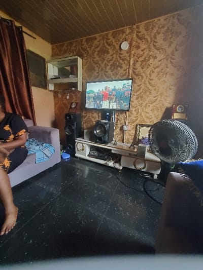 The image depicts an indoor living room in Isunba, Nigeria. A person, gender indeterminate, is seated on a light grey fabric couch in the foreground on the left, wearing a dark garment with orange patterns; their bare left foot is visible. A light blue and white patterned blanket is draped over the couch armrest next to the person.

A flat-screen television is mounted on the patterned golden-brown wallpapered wall, displaying an outdoor scene featuring multiple individuals. Below the television, a white and gold media console supports a central speaker, a gold-colored miniature Eiffel Tower replica, and various decorative items, including awards or plaques. Two black tower speakers flank the console; one is partially obscured by the couch on the left. A black oscillating stand fan is positioned to the right of the media console.

The walls are covered with golden-brown patterned wallpaper. The ceiling is constructed from light brown paneled material. The floor is composed of dark, glossy tiles with lighter speckles. A window with drawn brown curtains is visible on the far left. Above and to the left of the television, a white wall-mounted display cabinet with glass doors is present. A round white device, likely a smoke detector, is mounted high on the central wall. Electrical cabling is visible behind the media console and television. A dark-colored couch or armchair is partially visible on the far right.