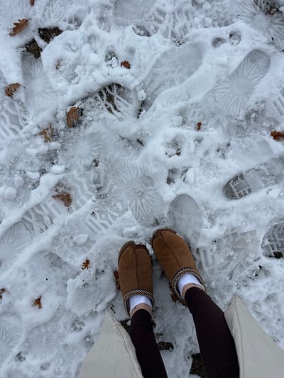 The image captures a downward-angled view of a snowy ground surface in Доња Брезна, Montenegro. The terrain is covered in a patchy layer of snow and ice, revealing areas of dark earth and sparse dry grass. Numerous distinct footprints are imprinted into the snow and ice, displaying various human shoe/boot sole patterns, including prominent radial and geometric tread designs, alongside smaller animal paw prints. Scattered across the snow and entangled within the exposed ground cover are several dry, brown oak leaves. In the lower center of the frame, the lower legs and feet of an individual are visible. The person wears dark brown leggings, white socks, and brown, moccasin-style boots featuring a patterned trim of white and brown around the ankle opening. A light cream or beige outer garment is partially visible on the lower right, adjacent to the right leg.