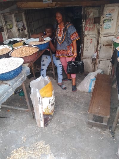 This image depicts a bustling scene at an outdoor market stall in Mgbuoshimini, Nigeria, focusing on two individuals and various grains. An adult woman, distinguished by her long dark hair (possibly dreadlocks or braids), stands on the right. She wears a grey dashiki-style tunic adorned with colorful patterns, pink capri-length pants, and dark footwear, carrying a black handbag. Her expression is a pleasant smile.

Beside her, a younger person, likely a boy or teenager, is actively engaged. He wears a dark sleeveless top and light blue jeans featuring prominent black 'CC' logos, paired with grey slide sandals. His hands are immersed in a large blue basin, seemingly scooping or sorting light-colored grains, possibly beans or rice. Several other large basins filled with different types of grains, including lighter and reddish varieties, are arranged around them, indicating a vendor specializing in staple food items.

The ground is concrete, with scattered grains visible, suggesting the ongoing activity of a market. A large white sack, partially open and clearly labeled "EMPIRE RICE," stands prominently in the foreground. Another smaller white sack and a rustic wooden bench are visible nearby. The background consists of weathered metal doors and panels, typical of market stalls, covered with various posters and some handwritten text, further establishing the informal market setting. The natural lighting suggests the scene takes place during the daytime. Both individuals appear comfortable and at ease, with the woman smiling, adding a warm touch to the vibrant market atmosphere.