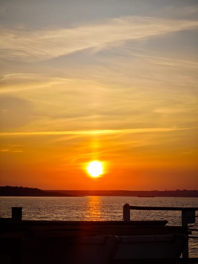 The image depicts a sunset over the Yenisei River in Igarka, Russia. The sun, a bright white orb, is positioned slightly above the horizon, casting a warm orange glow across the sky and reflecting on the water. Wispy clouds streak across the upper portion of the sky, transitioning from a pale yellow near the horizon to a muted grey at the top. In the foreground, the dark silhouette of a wooden pier or deck is visible, with a railing on the right side. A dark cylindrical object, possibly a bollard, stands on the left side of the pier. The far bank of the river is a dark, silhouetted landmass, with a faint outline of buildings on the horizon to the right. The water is calm, with gentle ripples reflecting the sunset. The overall scene is one of serene natural beauty during twilight.