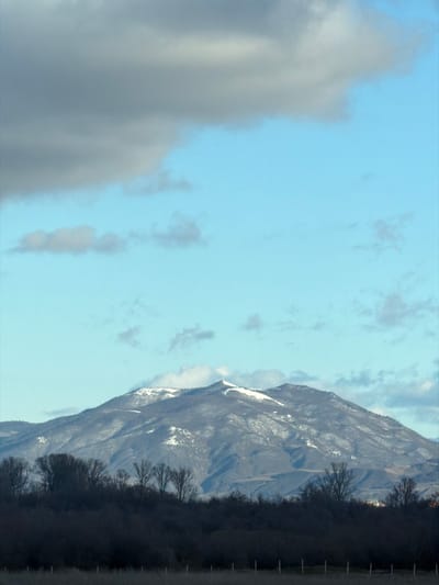 The image displays a wide-angle natural landscape captured in the vicinity of Akhalsopeli, Georgia. The upper 60% of the frame is occupied by a sky showing a large, dark grey cloud mass in the upper left, transitioning into clear, light blue sky with scattered smaller, lighter grey clouds towards the right and bottom. A prominent mountain range dominates the mid-ground, with its peaks and upper slopes covered in irregular patches of white snow against dark, un-vegetated sections. Below the mountains, a dense, horizontal band of dark, silhouetted deciduous trees, bare of leaves, spans the width of the image. The immediate foreground consists of a dark, shadowed field or scrubland, with a faint horizontal line of dark, slender vertical elements consistent with a fence line or sparse posts. No individuals or explicit human activities are present.