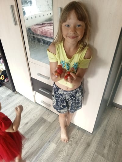 A young Caucasian girl with blonde hair and blue eyes, wearing a yellow off-the-shoulder top with a pineapple pattern and blue and white patterned shorts, stands in front of a light-colored wardrobe with a mirrored door reflecting a bed with a pink blanket. She is holding a few strawberries in her hands and smiling at the camera. Another child, out of focus and wearing a red dress, is partially visible in the lower left corner of the image. The floor is made of light gray wooden planks. The scene appears to be indoors, likely a bedroom or living space in a residential building in Buraly, Russia.