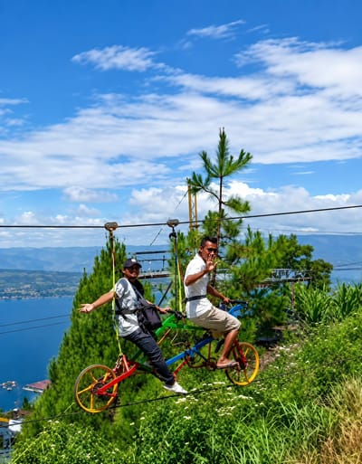 Two men are riding a tandem bicycle suspended from a zip line over a lush green hillside. The man in the front is wearing a black baseball cap, a gray t-shirt, dark pants, and white sneakers. He has a black backpack slung over his shoulder and is holding his left arm out to the side, with his palm facing outward. The man in the back is wearing a white t-shirt, light brown shorts, and flip-flops. He is waving at the camera with his right hand. The bicycle is colorful, with red and yellow wheels. In the background, a large body of water, likely a lake or ocean, is visible, with mountains and a city on the horizon. The sky is blue with scattered white clouds. The scene appears to be an adventure park or tourist attraction in a scenic location.