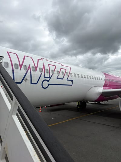 A large passenger airplane, specifically a Wizz Air aircraft, is prominently featured, parked on an airport tarmac under a heavily overcast sky. The airplane's white fuselage, adorned with the distinctive pink and blue "WIZZ air" logo, occupies the central and left portions of the frame. Numerous oval windows are visible along the side of the aircraft.

In the foreground, a white and dark grey railing, likely from a mobile staircase or passenger boarding bridge, cuts diagonally across the bottom right. On the tarmac, a yellow line marks a boundary, and a person wearing high-visibility clothing is visible near the plane's underside, suggesting ground operations are in progress. Further back, part of an engine or wing and ground equipment, including what appears to be the front of a service vehicle and orange safety cones, are seen near the aircraft's rear.

The sky is dominated by dark, heavy clouds, indicating an overcast or potentially stormy day. The scene depicts an airport apron, with the aircraft seemingly awaiting departure or having recently arrived at an airport in Tillé, France.
