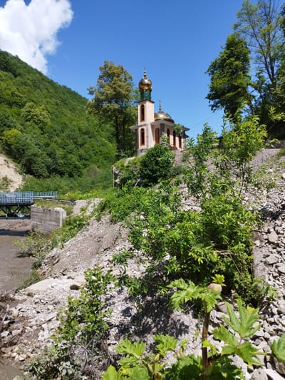 The image captures a mosque situated on a hillside in Kiri, Russia, under a clear blue sky. The mosque features two golden domes and a tall minaret. In the foreground, lush green vegetation grows amongst rocks, with a muddy river flowing to the left, spanned by a blue-railed bridge supported by concrete pillars. The hillside behind the mosque is densely covered with green trees. The scene is bright and appears to be taken on a sunny day.